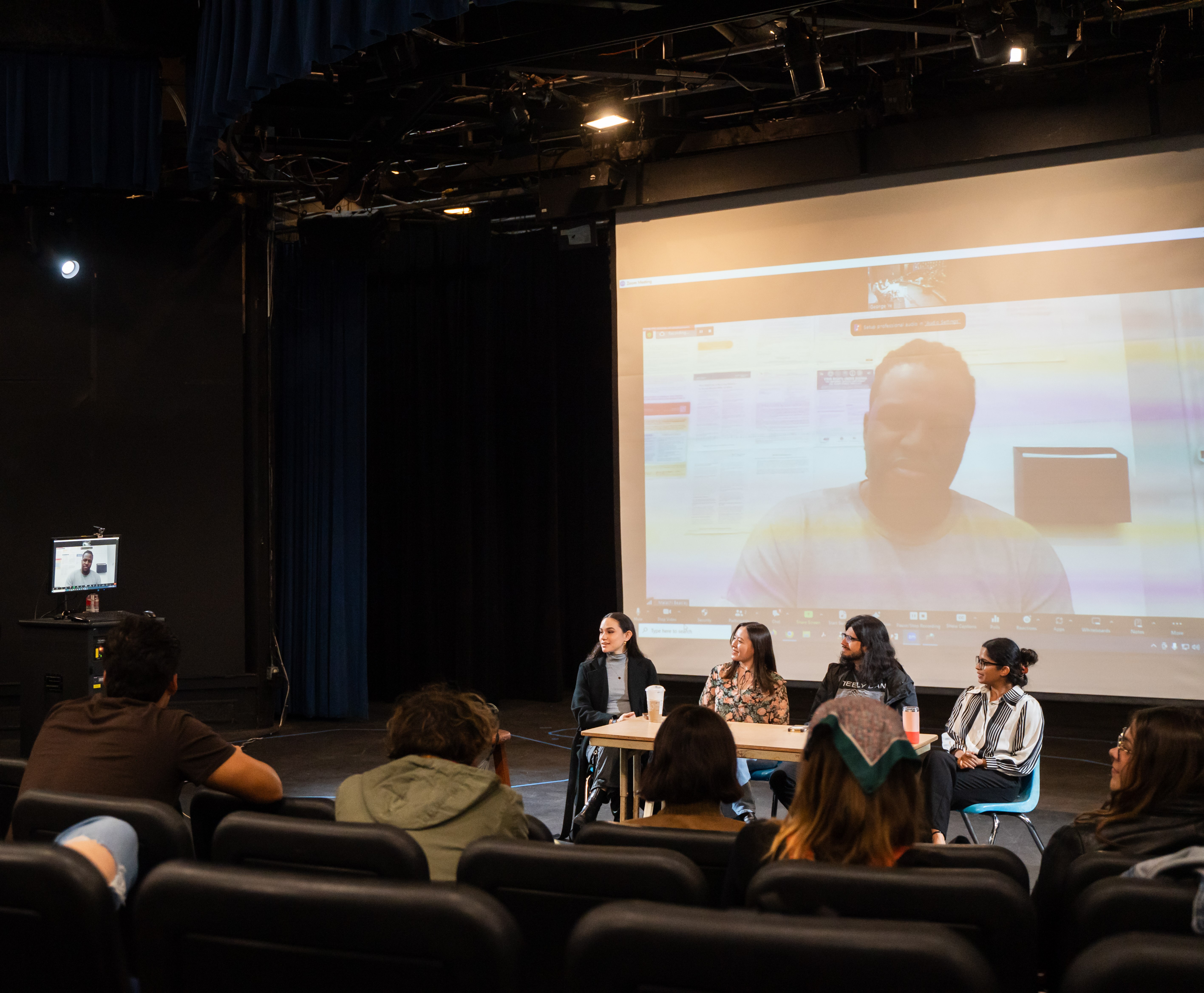 students watch panel of alumni, 4 alumni at table with one on large screen behind them, speaking via zoom